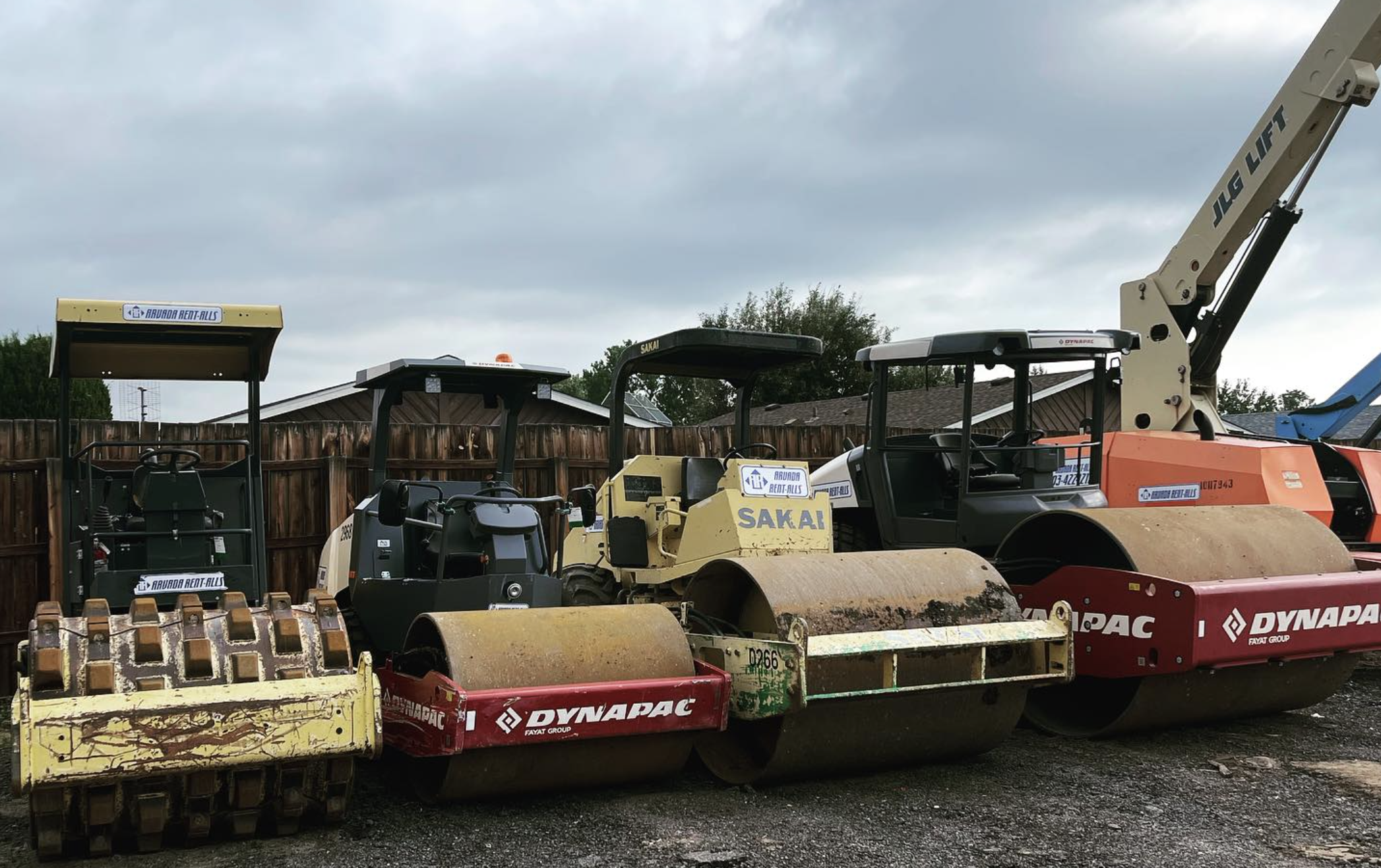 Row of heavy construction equipment, including several Dynapac and Sakai road rollers and a JLG telehandler, parked side-by-side in a gravel lot under a cloudy sky.