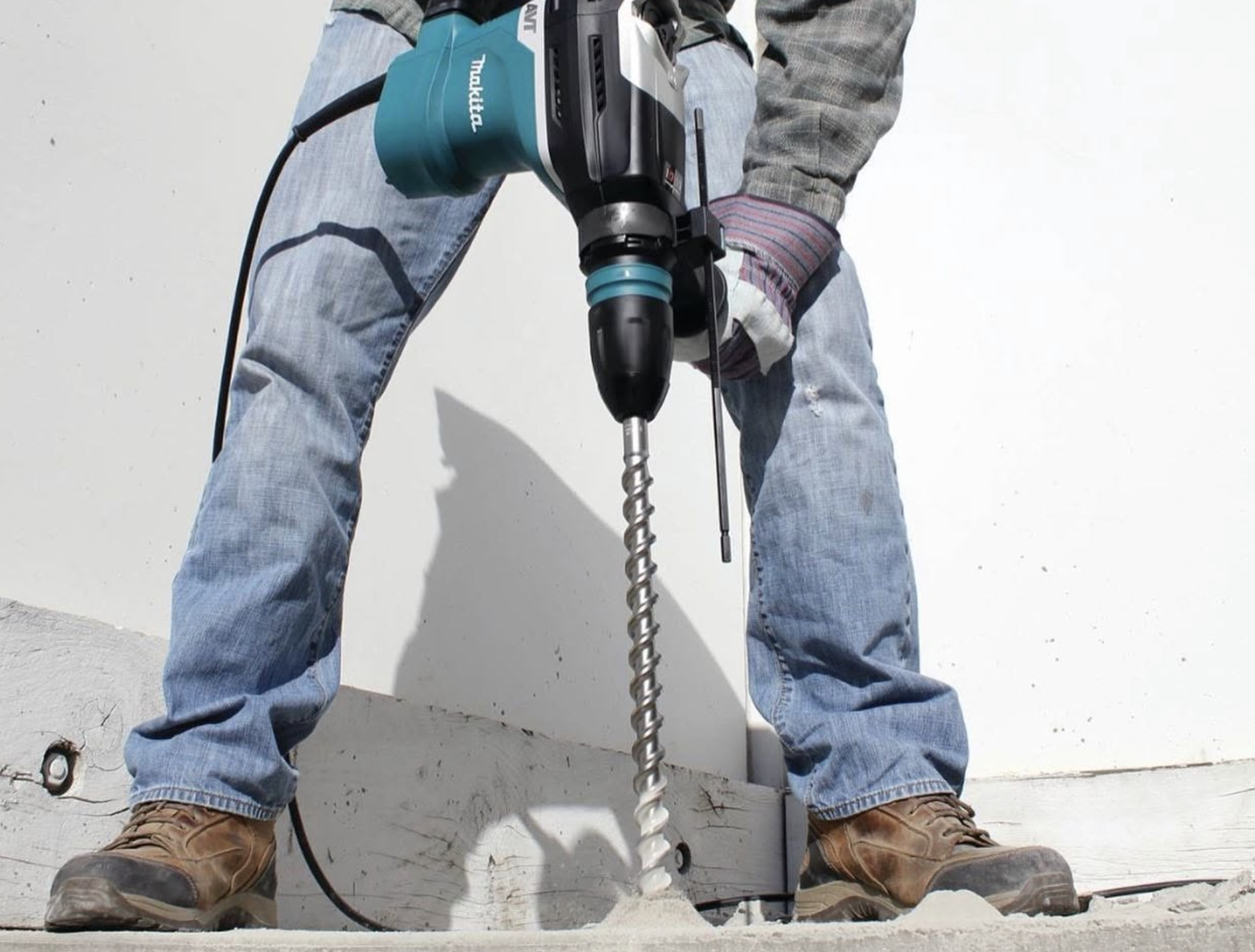 Person operating a Makita rotary hammer drill with a large auger bit, drilling into a concrete slab while wearing work gloves, jeans, and brown work boots.