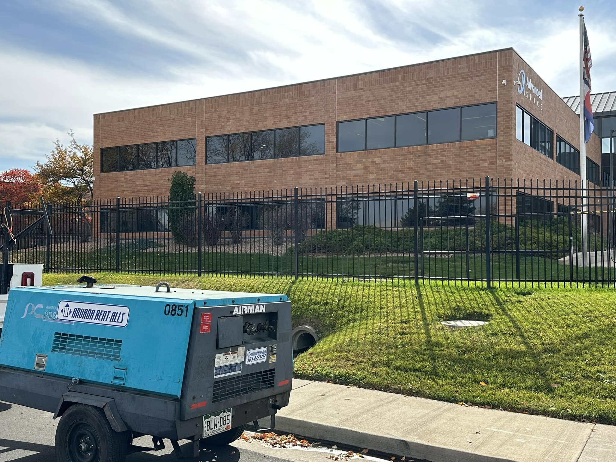 A two-story brick office building labeled 'Advanced Space' behind a black iron security fence, with a teal Airman portable air compressor from Arvada Rent-Alls parked on the street in the foreground.