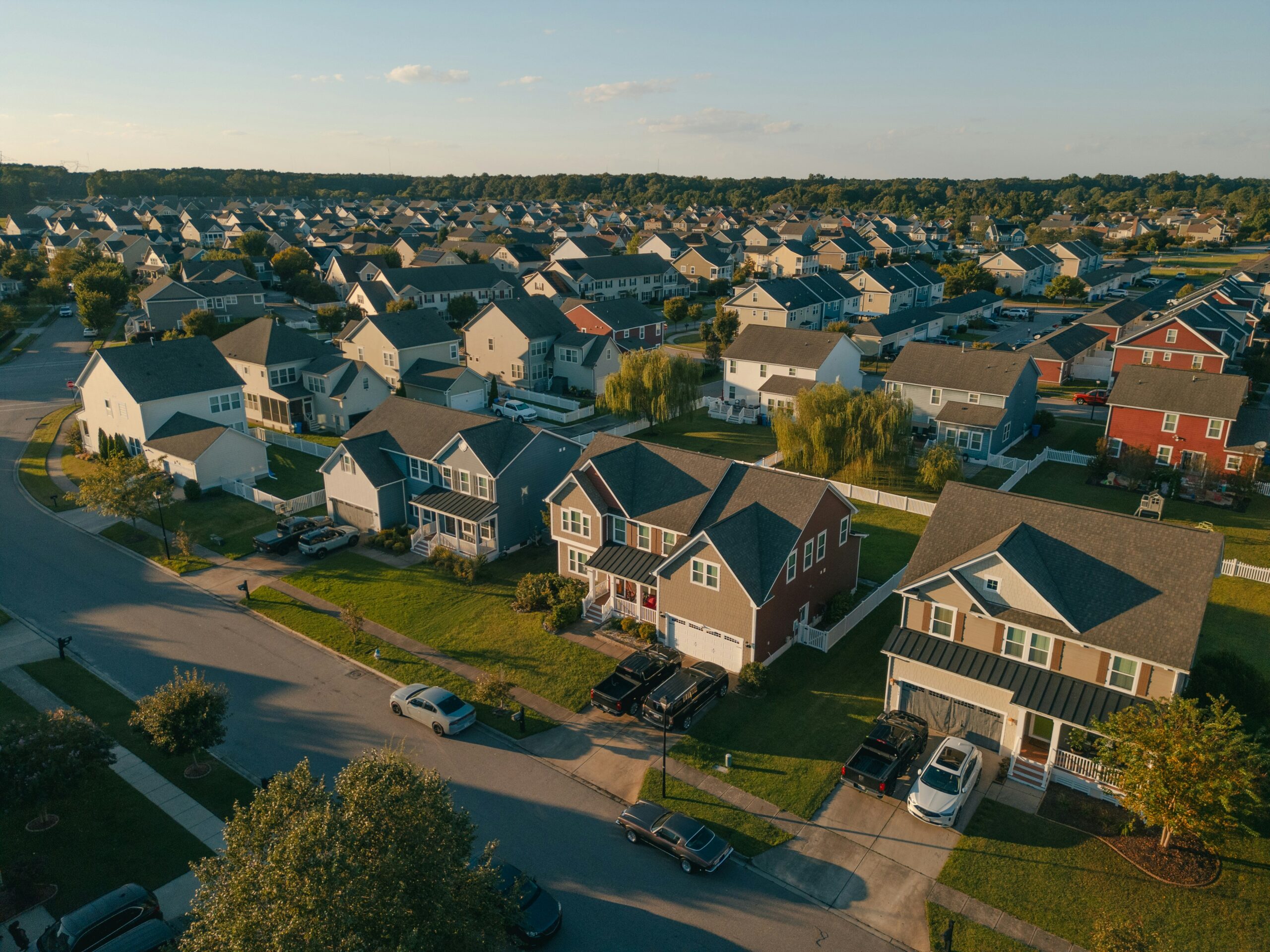 Aerial view of a suburban neighborhood at golden hour, with rows of two-story houses, driveways, and tree-lined streets bathed in warm evening light.